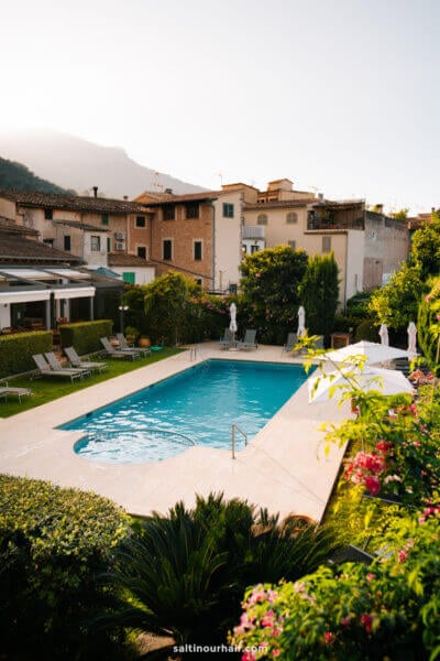 Rectangular outdoor swimming pool at Hotel Soller, Mallorca, Balearic Islands, surrounded by lounge chairs and greenery, with authentic buildings and mountains in the background under a clear sky.