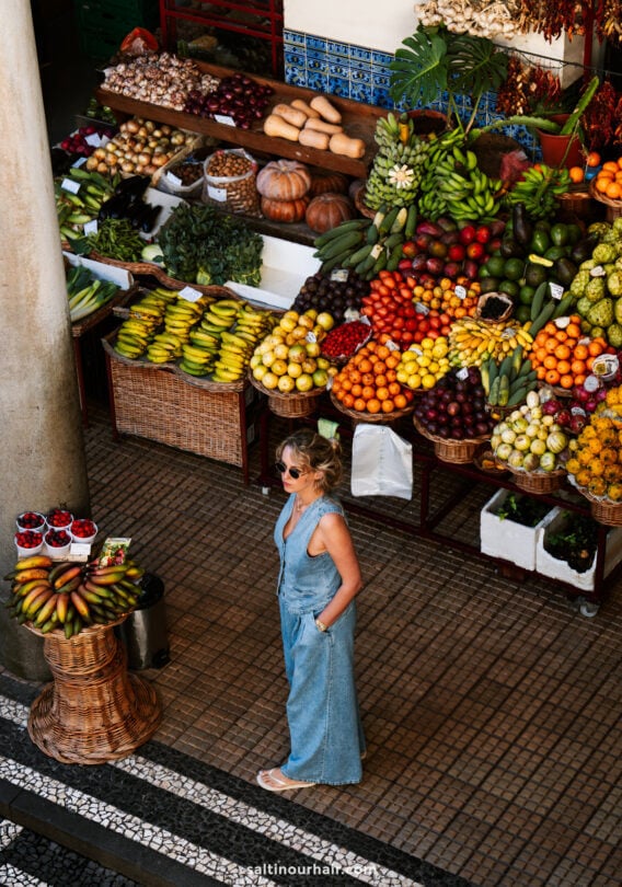 A woman wearing sunglasses and a blue jumpsuit stands in front of a vibrant market in funchal Madeira stall, surrounded by an array of colorful fruits and vegetables.