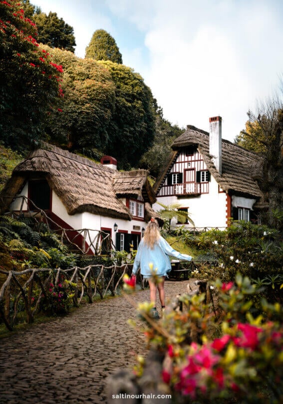 A woman in a blue shirt walks on a stone path toward traditional thatched-roof cottages, surrounded by lush greenery and flowers—evoking the charm of both Madeira vs Azores landscapes.
