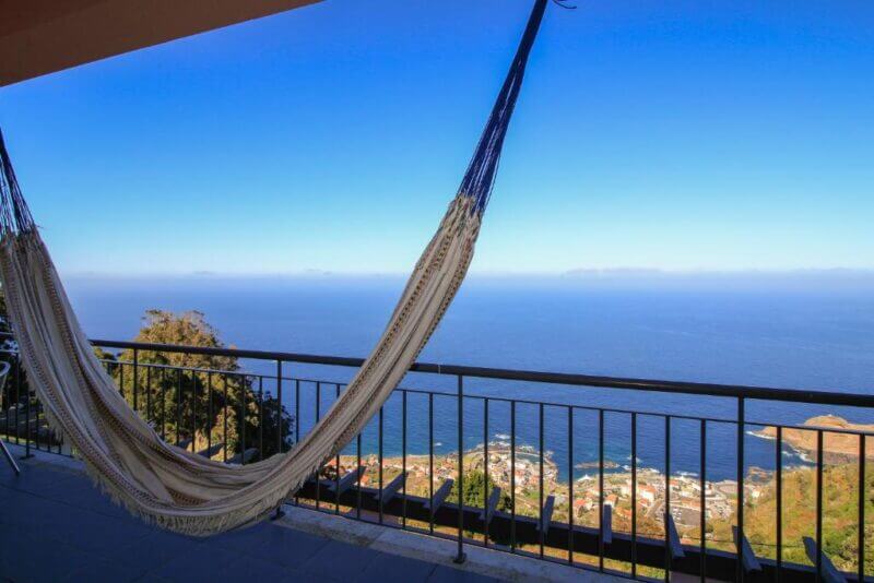 A hammock hangs on a balcony overlooking the Paradise Ocean View of Porto Moniz, with a wide expanse of blue ocean and the coastal town below under a clear sky.