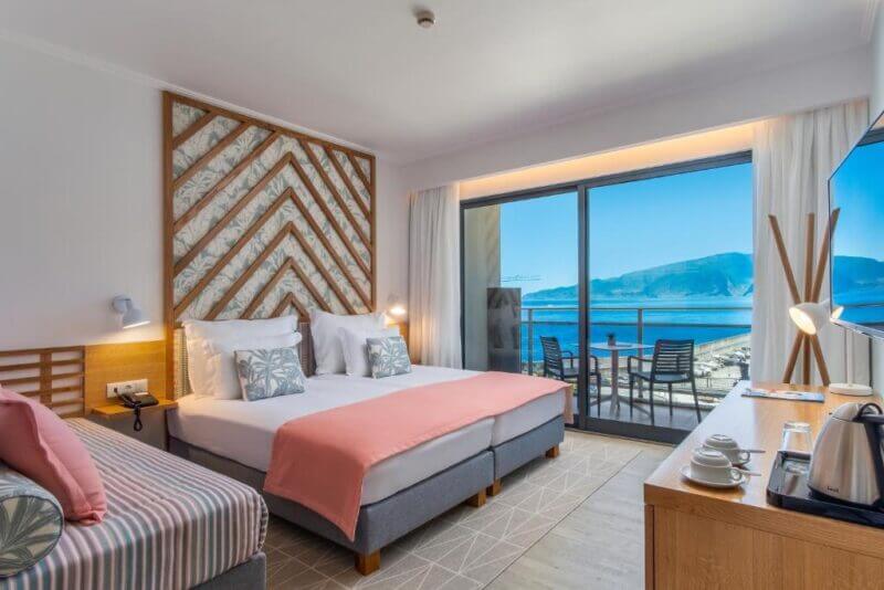 A modern hotel room at Aqua Natura Bay features two beds, a sofa, a balcony with chairs, and stunning views of the sea and mountains through large sliding glass doors in Porto Moniz.