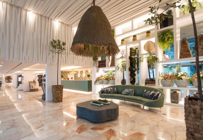 Hotel Alto Lido's lobby in Funchal features marble floors, a modern green sofa, a large hanging lamp, indoor plants, and illuminated display shelves adorned with decorative items and flowers.