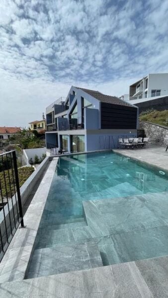 Modern two-story house with large windows and balconies, overlooking a rectangular outdoor swimming pool in an Azul hue, under a partly cloudy sky reminiscent of Funchal’s tranquil ambiance.