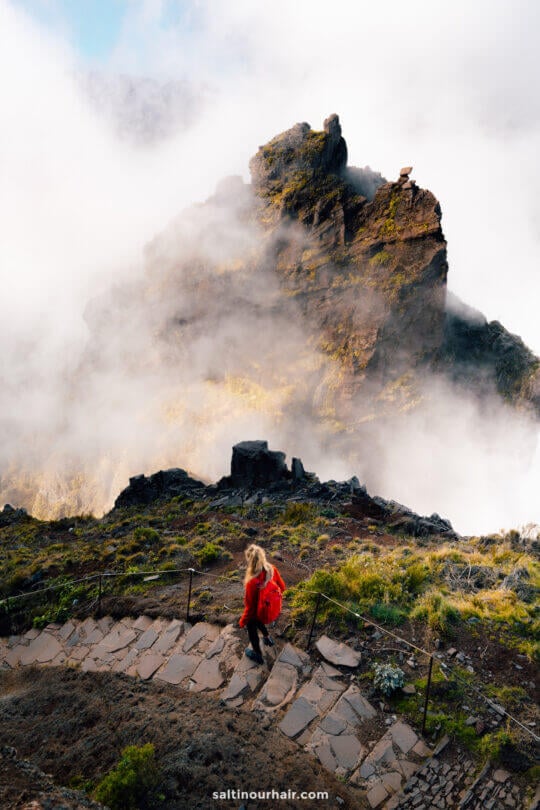 A person with a red backpack walks down a stone path on a mountain, surrounded by mist and rocky peaks, enjoying the scenic PR1 hike in Madeira.