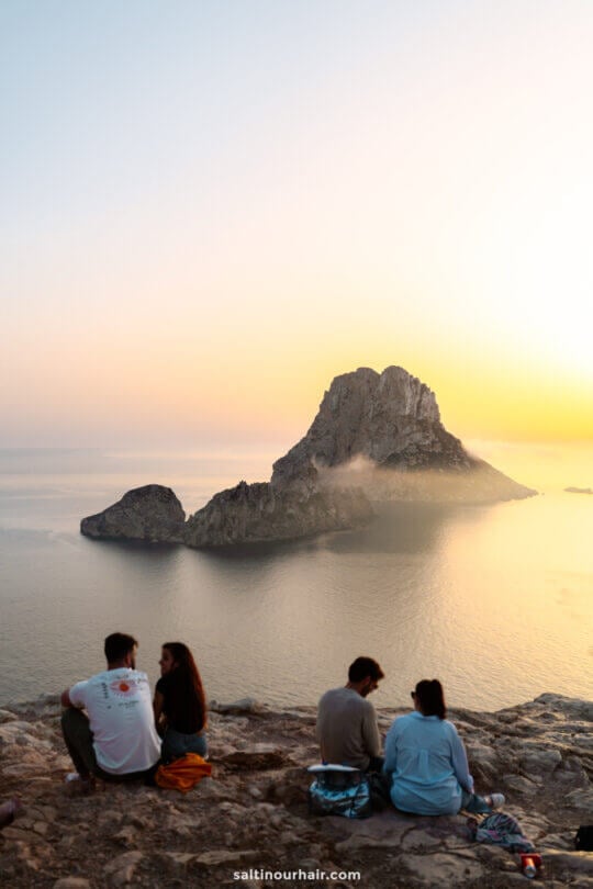 Four people sit on a rocky cliff overlooking the sea at sunset, gazing at the iconic Es Vedra island off the coast of Ibiza in the Balearic Islands, with calm water and a dramatic landscape in the background.