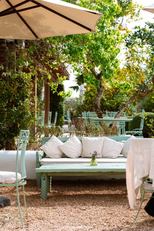 Outdoor patio inspired by Ibiza, with a white cushioned bench, small table with a vase of flowers, mint green chairs, large umbrellas, and lush greeneryâ€”evoking the relaxed charm of the Balearic Islands.