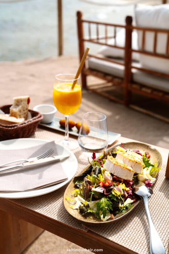Outdoor table setting at a restaurant on the beach in Ibiza, featuring a plate of salad topped with goat cheese, a glass of orange juice, bread basket, and cutlery on a napkin.