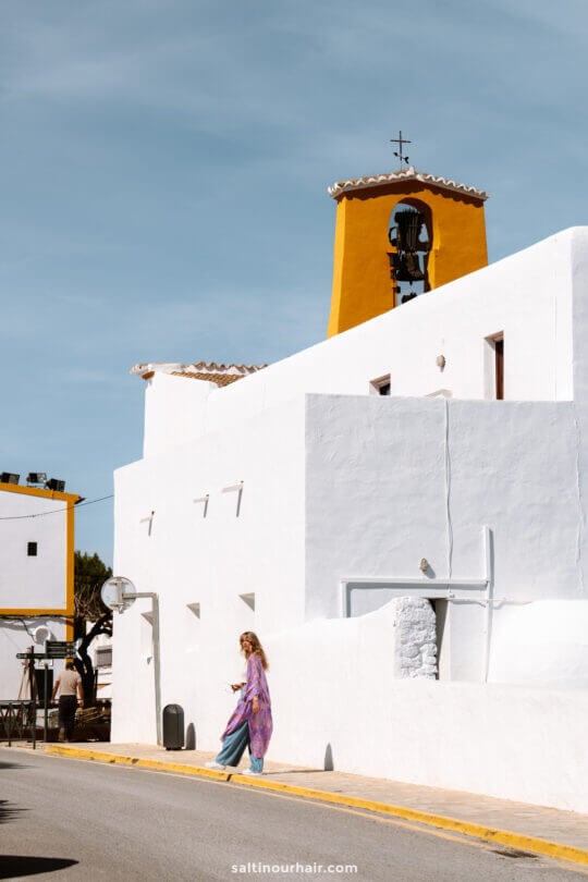 A woman in colorful clothing walks past a white building with yellow trim and a bell tower under a clear blue sky in Ibiza, Balearic Islands.