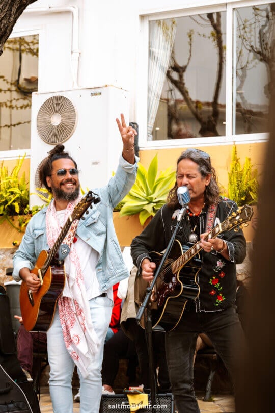 Two men play acoustic guitars and sing outdoors in Ibiza, Balearic Islands; one raises his hand in a peace sign.