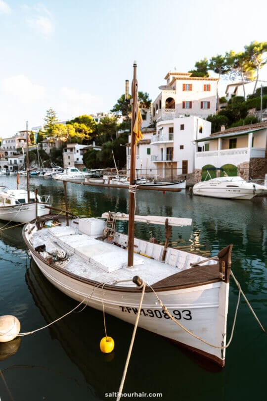 A small wooden boat is moored in a calm harbor with white houses and green trees on the hillsideâ€”a tranquil scene that captures one of the many picturesque things to do in Mallorca.