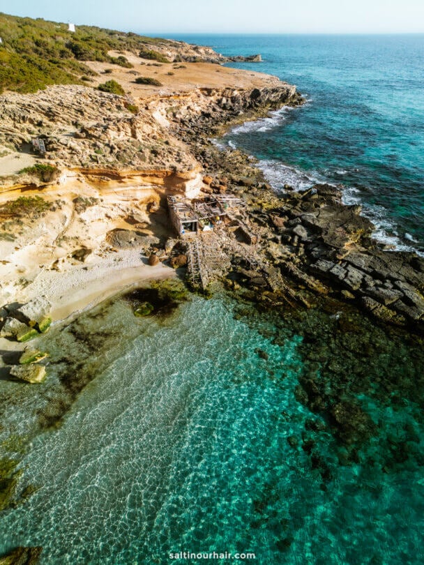Aerial view of a beach in Formentera, Balearic Islands, shows a rocky coastline with clear turquoise water and a small stone structure near the shore, surrounded by sparse vegetation.