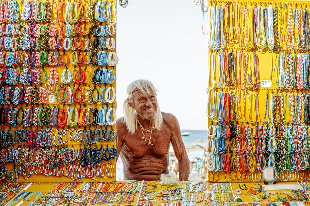 A shirtless older man with long white hair stands in a colorful jewelry stall in Formentera, Balearic Islands, displaying various beaded necklaces and bracelets on a yellow background.