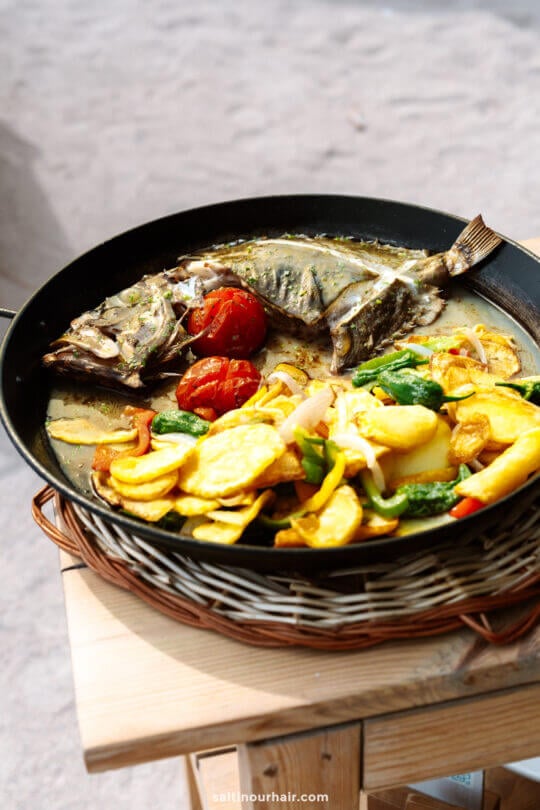 A cooked whole fish, inspired by the Balearic Islands, served in a pan with roasted tomatoes, potatoes, and mixed vegetables on a wooden table.