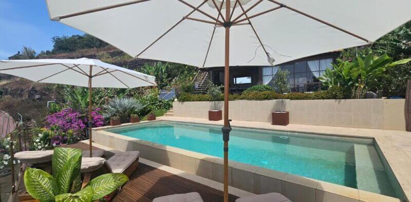Rectangular pool with clear water, surrounded by tan stone deck, lounge chairs, and large white umbrellas; landscaped garden and modern Suites Calheta house in the background.