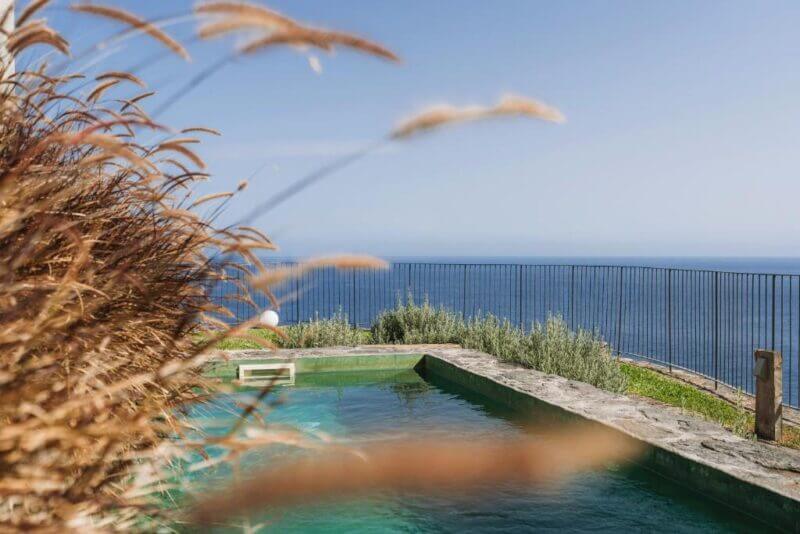 A swimming pool with clear water at Socalco Nature overlooks the ocean in Calheta, bordered by a metal fence and tall grasses on a sunny day.