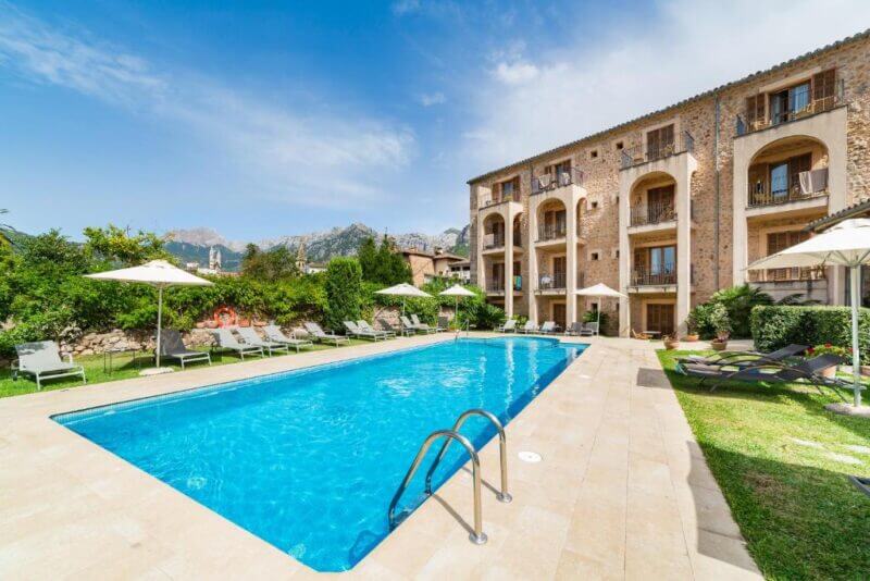 A rectangular outdoor swimming pool with lounge chairs and umbrellas beside the multi-story stone hotel Ca'l Bisbe in Soller, set under a clear blue sky.