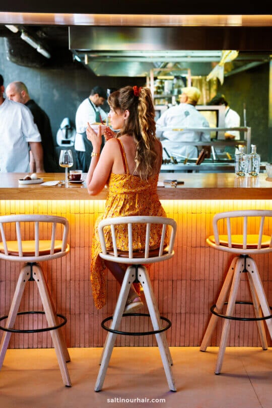 A woman in a yellow dress sits at a bar counter, holding a drink, with chefs working in the background—a stylish scene capturing the lively dining atmosphere among the top things to do in Funchal, madeira