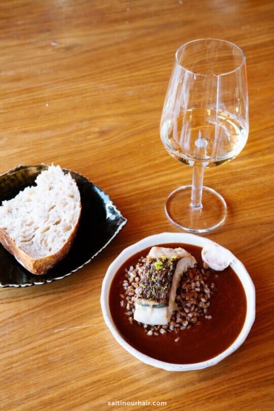 A plate with fish on grains in sauce, a slice of bread on a side dish, and a glass of white wine on a wooden table—a perfect way to enjoy local flavors while exploring things to do in Funchal, madeira