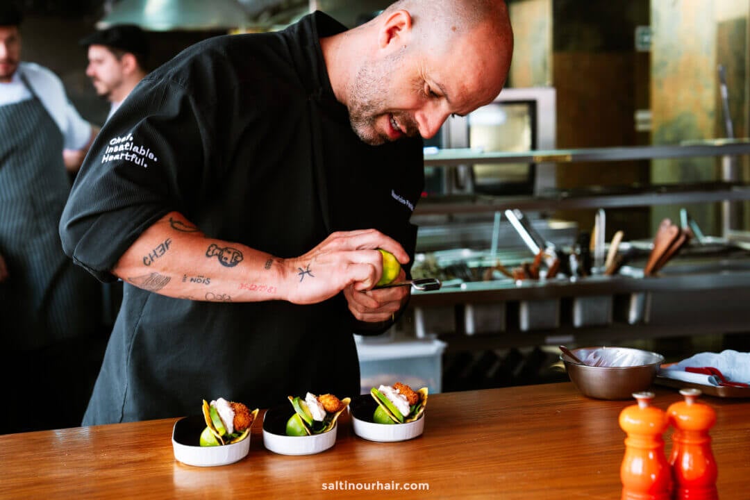A chef garnishes small dishes of food in a professional kitchen in Funchal, with kitchen utensils and staff visible in the background—perfect for anyone exploring things to do in Funchal, madeira