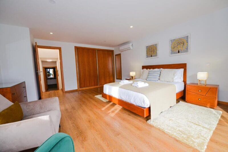 Spacious bedroom at Casa do Cantinho with a double bed, wooden furniture, two nightstands with lamps, a beige armchair, and large built-in wardrobes. The floor is hardwood, and artwork decorates the walls in this Sao Vicente retreat.