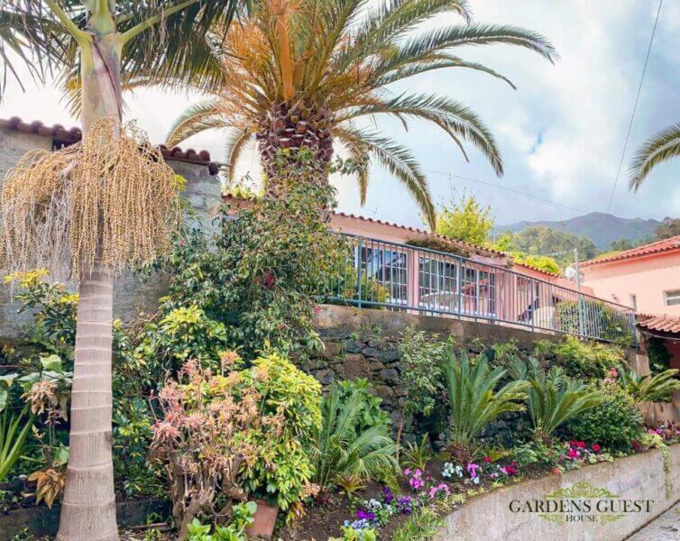 A lush garden with tropical plants and palm trees welcomes you to Gardens Guest House, a pink home with a red-tiled roof and metal railing in Sao Vicente, with mountains visible in the background.