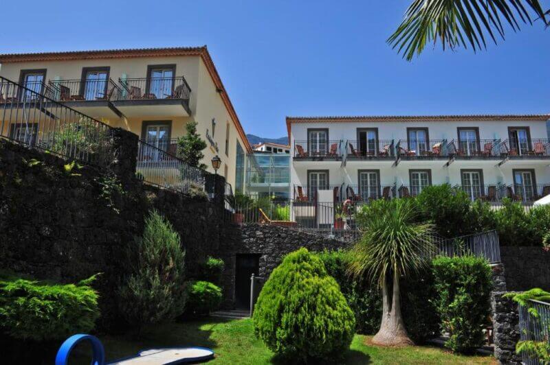 Two three-story buildings with balconies overlook a landscaped garden with shrubs, trees, and stone walls at Estalagem do Vale hotel in sunny Sao Vicente.