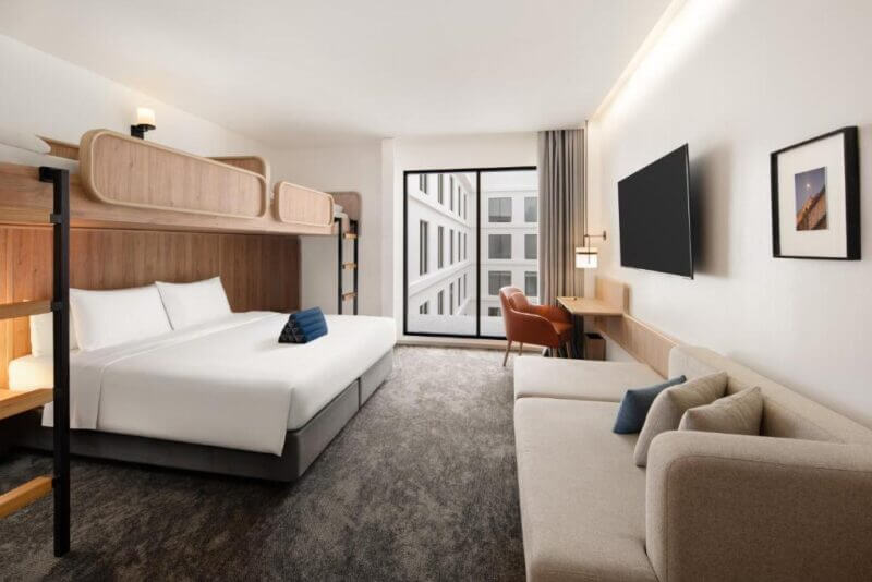 Modern ASAI Bangkok hotel room with a large bed, upper bunk, sofa, desk, chair, wall-mounted TV, and window overlooking Bangkok Chinatown’s vibrant streets.