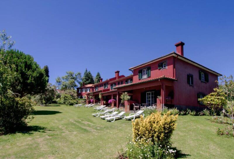 A row of red, two-story houses at Quinta Santo Antonio with chimneys and green shutters sits on a grassy lawn with lounge chairs under a clear blue sky.
