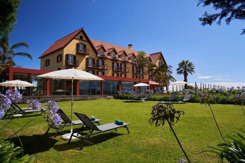 A well-maintained lawn with lounge chairs and umbrellas in front of the large, multi-story Hotel Quinta Do Furao, set in scenic Santana under a clear blue sky.