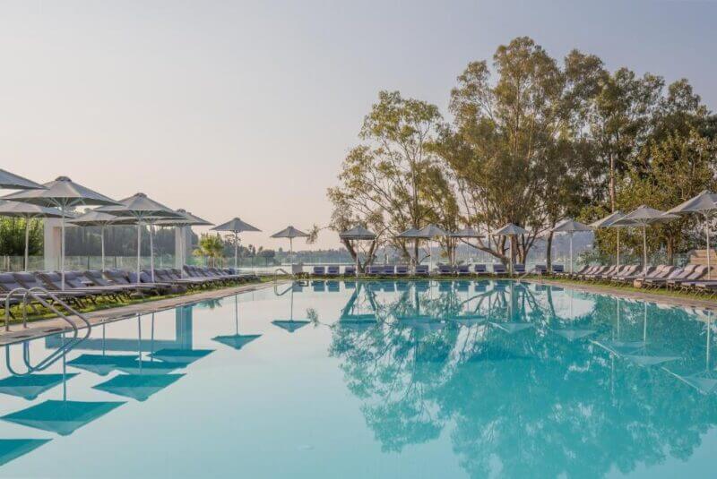 Outdoor swimming pool with clear water at Rodostamo Hotel, surrounded by lounge chairs and white umbrellas, trees in the background, under a clear sky—your perfect Corfu hotel escape.