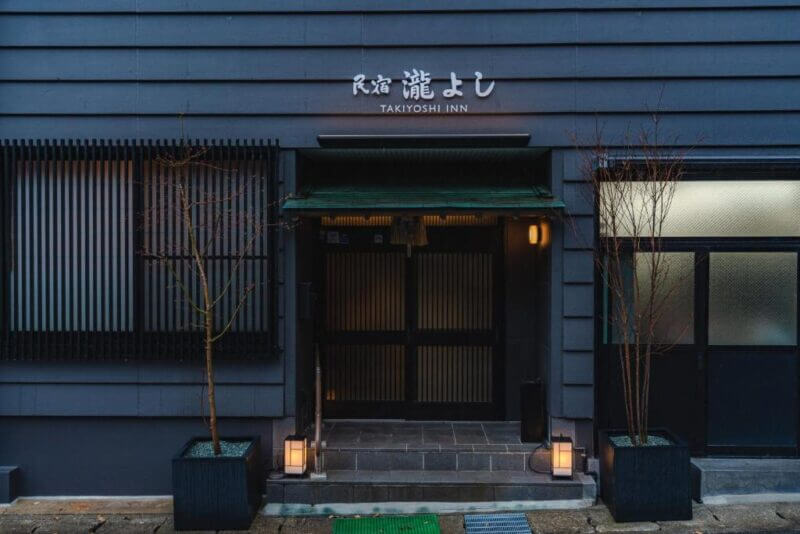 Exterior of Takiyoshi Guest House in Yunomine, featuring a dark grey facade, potted plants flanking the entrance, and glowing lanterns by the door.