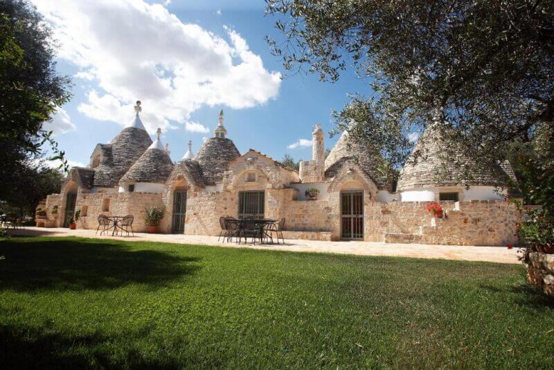 Trulli Boccadoro is a stone building with multiple conical roofs, outdoor tables and chairs, and a green lawn, set under a partly cloudy sky in scenic Ostuni.