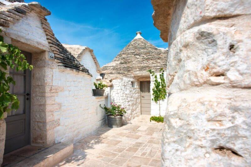 Stone buildings with conical roofs and white walls under a blue sky, with plants and a paved walkway in the foreground—perfect for a memorable stay at Trulli Holiday in Alberobello.