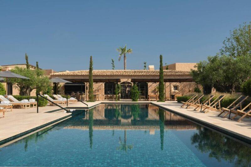 Rectangular outdoor swimming pool at Can Ferrereta in Santanyi, surrounded by lounge chairs, umbrellas, and lush greenery, with a stone building in the background under a clear blue sky.
