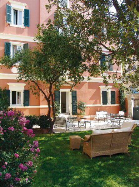The pink Siora Vittoria Boutique Hotel in Corfu features green shutters and overlooks a garden with a wicker sofa, metal chairs, tables, and flowering shrubs under leafy trees.