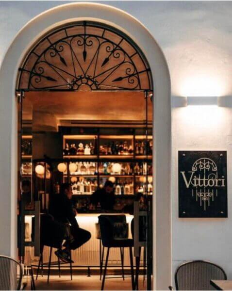 Bar interior with two people seated at the counter, viewed through an arched doorway. The sign outside reads “Vittori Corfu Rooms.” Shelves with bottles line the background, capturing the charm of Corfu accommodation.