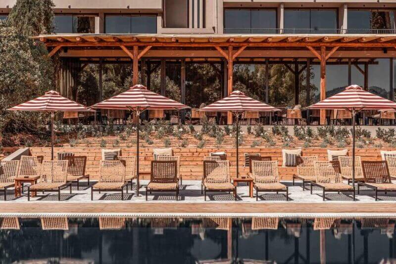 Outdoor pool area at Cook's Club Corfu Hotel, featuring wicker lounge chairs, red-and-white striped umbrellas, and a wooden terrace in front of a building with large windows.