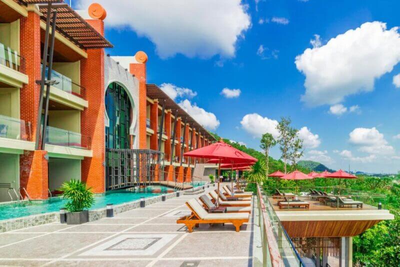 Modern hotel resort with a pool, sun loungers, and red umbrellas on a sunny day. Enjoy stunning blue skies and scattered clouds at Phu Pi Maan in Aonang.