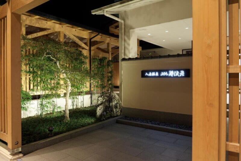 Entrance to Iruka Onsen at night, a modern Japanese building with wooden structures, a lit sign on the wall, and a landscaped garden with trees and rocks beside Hotel Kumano.
