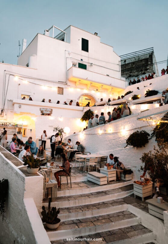 Outdoor restaurant where people dine and socialize on stone steps—capturing the lively, relaxed spirit of Ostuni in Italy—an idyllic moment on any Puglia itinerary.