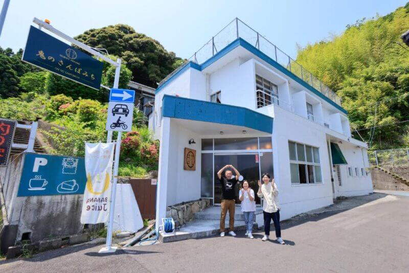 Three people stand in front of a white two-story building with a blue roof on a sunny day along Oninosanpomichi in Kumano. Signs and greenery are visible nearby.