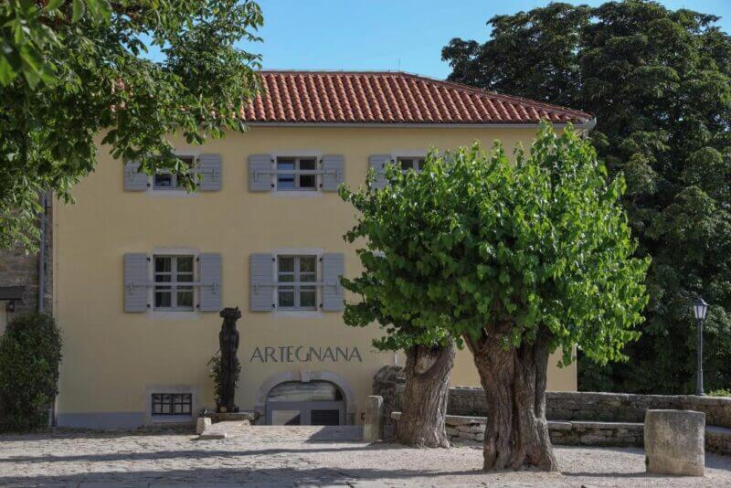 A yellow building with red roof tiles and gray shutters, labeled BB Artegnana 1798, stands behind two large trees in a sunlit stone courtyard in Groznjan.