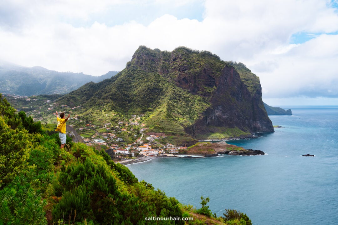 Een man staat op een groene heuvel bij een van de mooiste uitkijkpunten die Madeira: Miradouro do BoqueirÃ£o. Hij kijkt uit over een kustdorpje, steile kliffen en de oceaan onder een gedeeltelijk bewolkte hemel.
