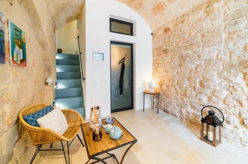 A cozy stone-walled entryway at B&B Monopoli features a wicker chair, a small table with decorative items, a lamp, and stairs leading up next to a black-framed glass door at this charming bed and breakfast.