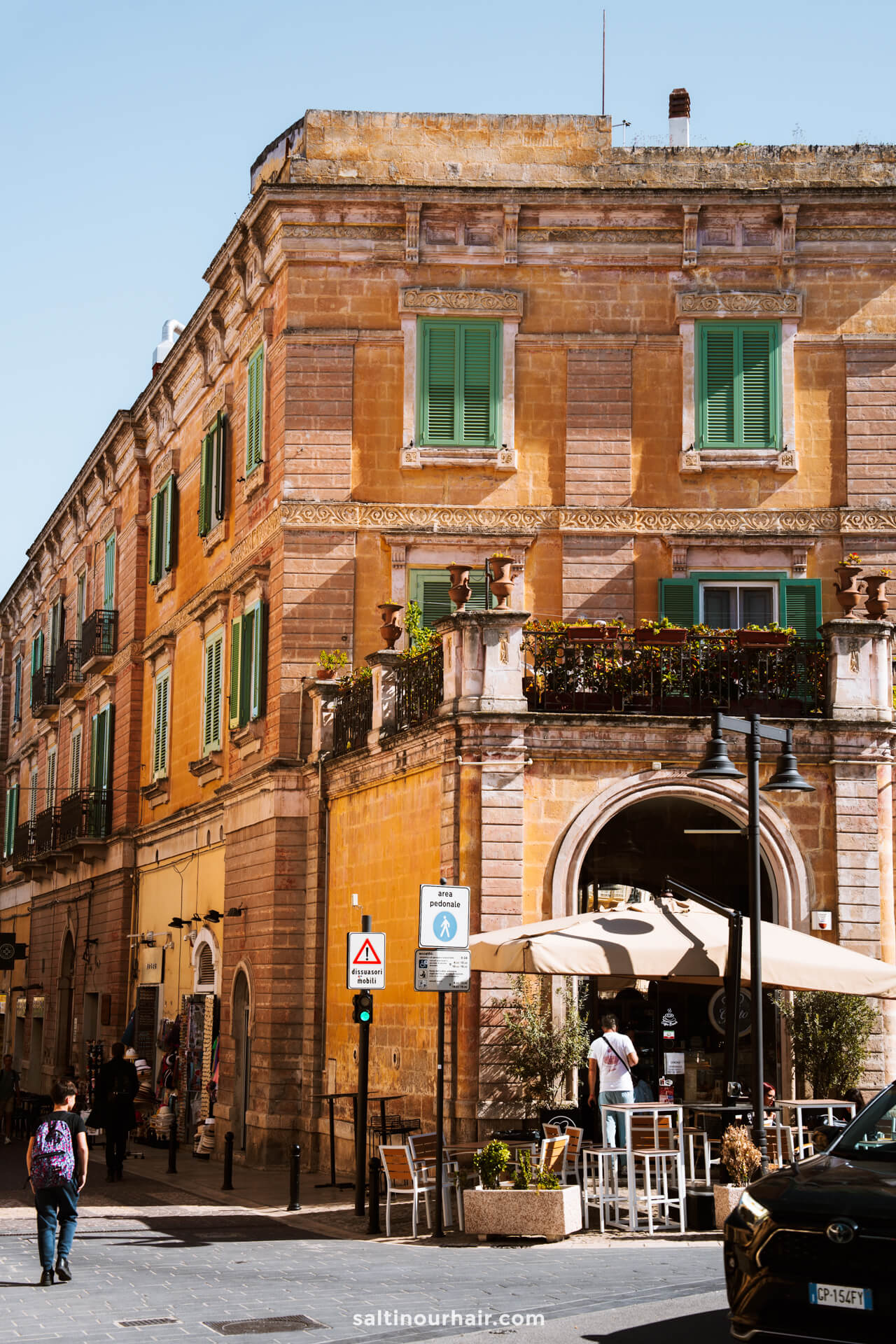 A historic, yellow-orange stone building with green shutters and a ground-floor café invites you to relax, as people stroll by on a sunny day—a charming scene perfect for any Matera Puglia itinerary.