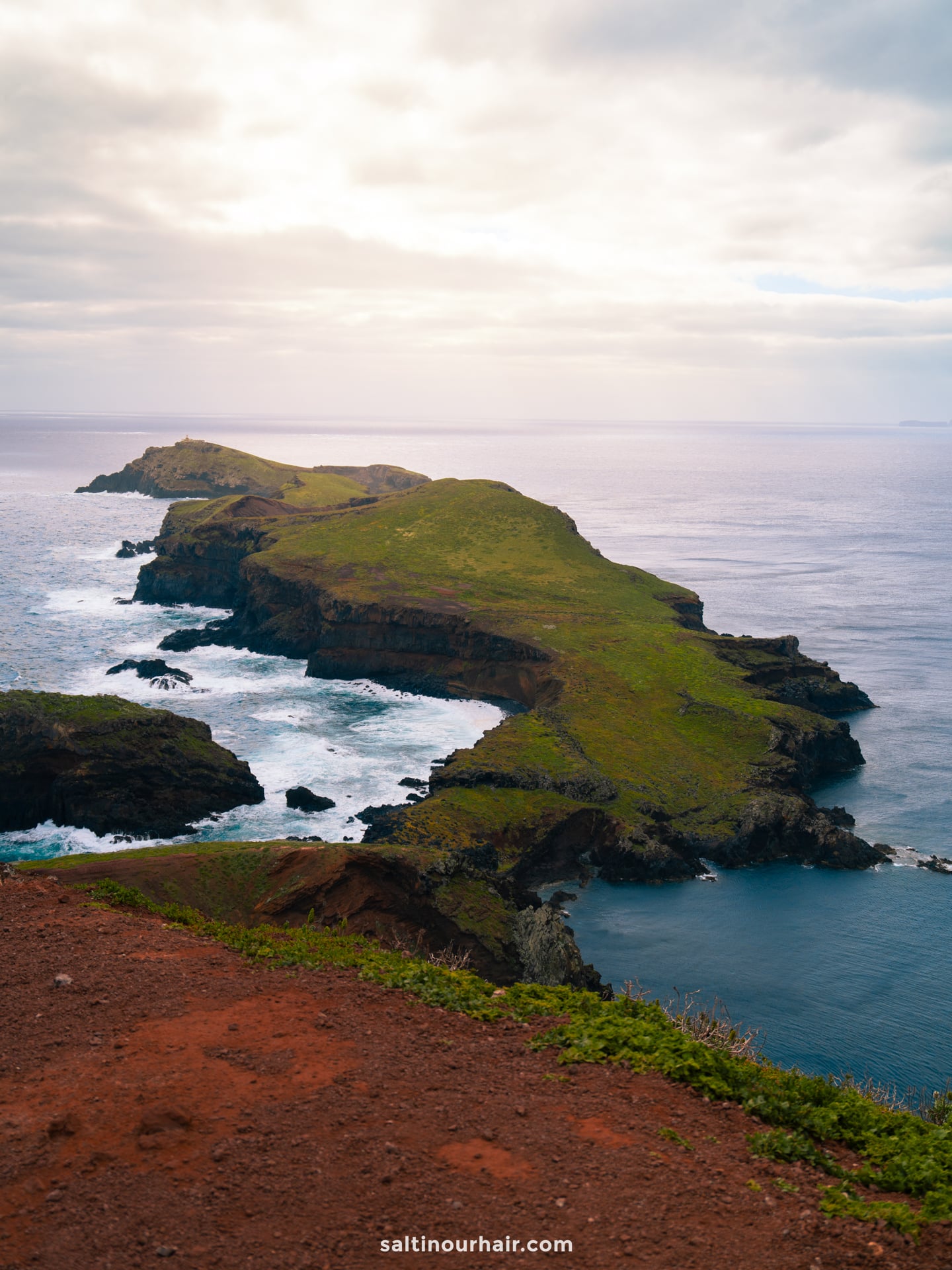 A rugged peninsula covered in green grass extends into the ocean, with waves crashing against its rocky cliffs under a cloudy sky—one of the best hikes Madeira has to offer.