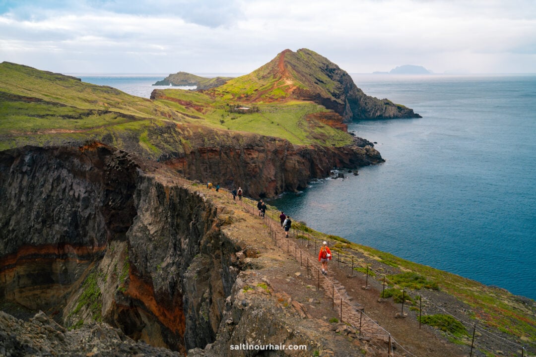 Hikers walk along a narrow, rocky coastal trail called Ponta do São Lourenço Hike — PR8 —one of the best hikes Madeira offers—with steep cliffs and green hills overlooking the ocean under a partly cloudy sky.
