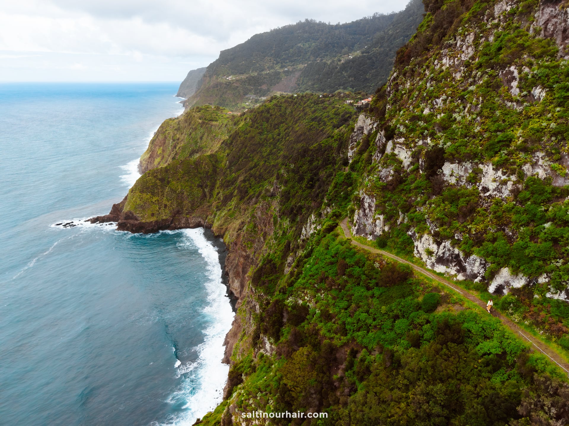 A narrow hiking trail runs along steep green cliffs overlooking the ocean, with waves crashing against the rocky shoreline below—one of the best hikes Madeira has to offer.