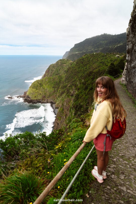 A woman with a red backpack stands on a stone path along a steep coastal cliff, overlooking the ocean and green hills under a cloudy sky—one of the best hikes Madeira has to offer.