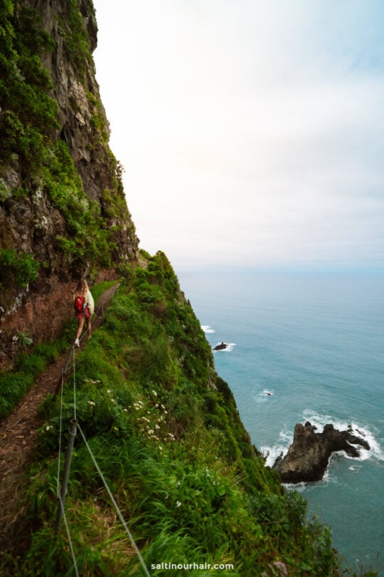 Person walking along a narrow trail on a steep, green cliff above the ocean—cloudy sky overhead and rocky outcrops below—captures the breathtaking scenery of one of the best hikes Madeira has to offer.